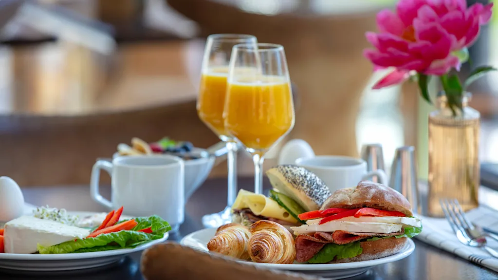 En frokostoppdekning med appelsinjuice, kaffe, bakverk, og en sandwich, pent arrangert på et bord med en rosa blomst i bakgrunnen.