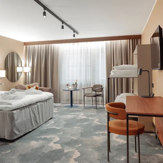 Ett prydligt arrangerat hotellrum har en stor säng med vita sängkläder, en våningssäng, ett skrivbord med lampa, ett bord med stol och mjukt ljus genom gardiner.