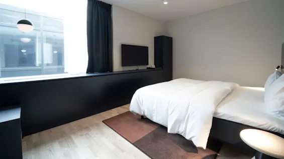 A neatly made bed with white bedding sits on a brown rug in a minimalist bedroom. A wall-mounted TV and large window with dark curtains provide a modern atmosphere.