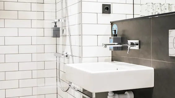 A white sink and toilet sit against tiled walls; adjacent is a glass-enclosed shower. A small shelf above the sink holds several glasses.