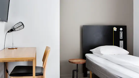 A single bed with white linens and a flower design on the headboard, alongside a wooden desk, chair, and modern lamp, in a minimalist room with a wall-mounted TV.