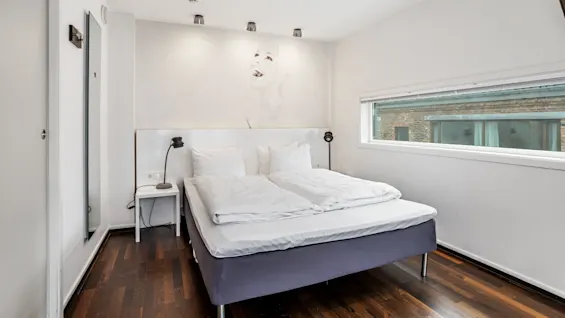 A neatly made bed stands in a bright room with artwork, wood flooring, and a large window overlooking a brick building.