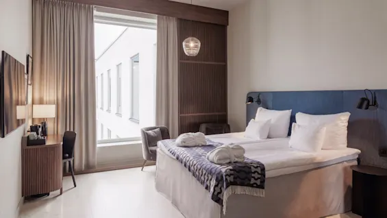 Neatly arranged hotel room with a made bed, side table, and chair, illuminated by natural daylight from a large window.