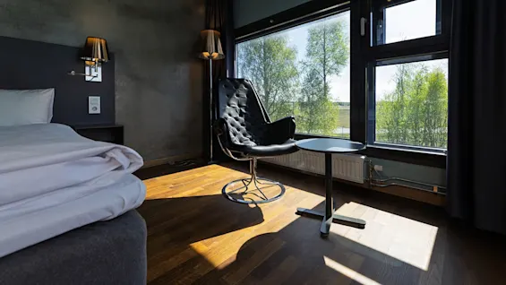 A black, modern chair faces a large window, letting in sunlight on hardwood floor. Nearby, a small round table and a neatly made bed create a cozy, minimalist room.