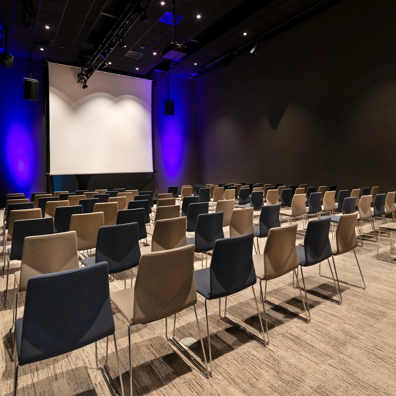 Rader med beige og blå stoler vender mot en stor projektor i et svakt opplyst konferanserom med blå lysaksenter mot veggene.