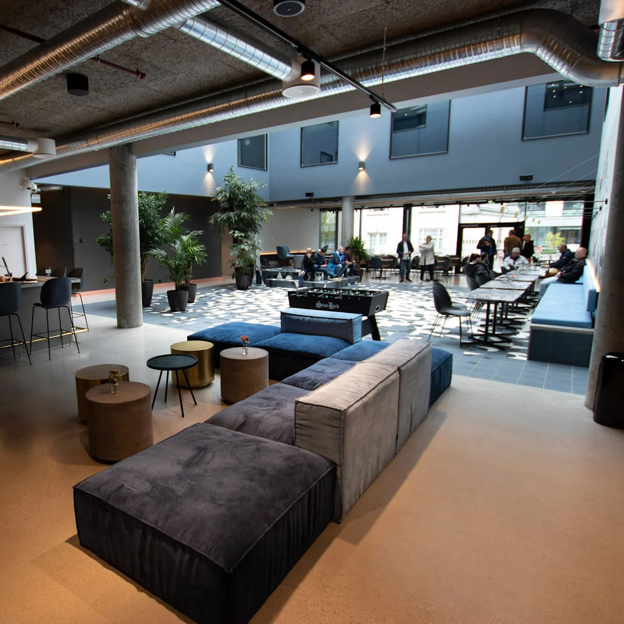 Rummelig lounge med moderne møbler, herunder sofaer og borde, og planter. Folk sidder rundt om et sort bordfodboldbord. Lys med højt til loftet og store vinduer.