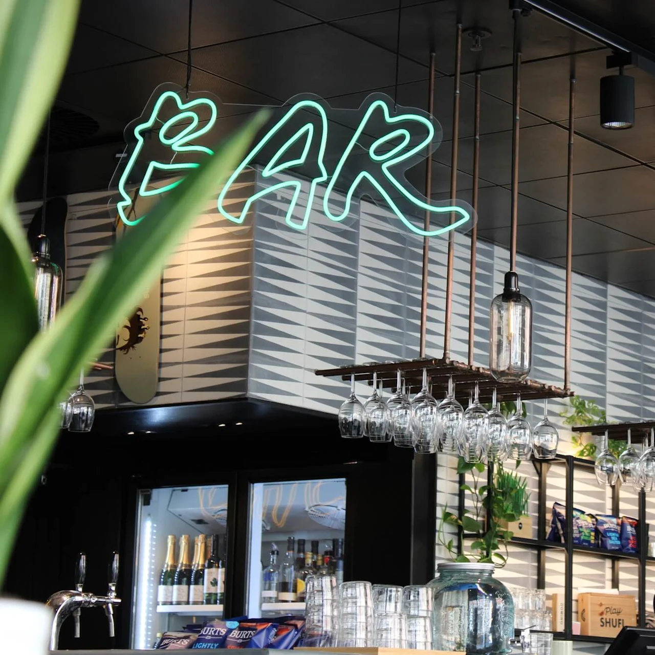 A modern bar with a hanging system for wine glasses and decorative lamps in an industrial style. Above the bar, a green neon sign displays the word "BAR."