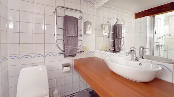 A bathroom with white tiles, wooden countertop, sink, mirror, towel rack, and shower.
