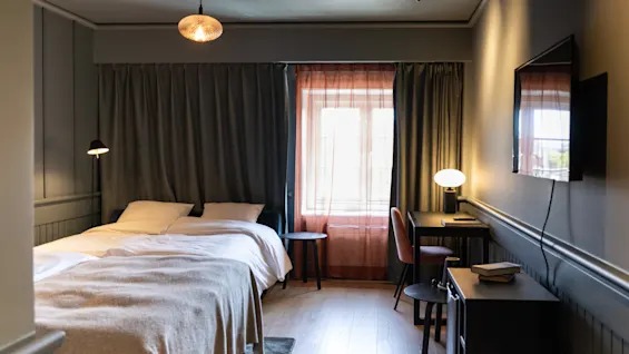 En pænt redt seng vender mod et vægmonteret TV i et blødt oplyst soveværelse. Et skrivebord med en stol og lampe er placeret ved et gardinrullet vindue.