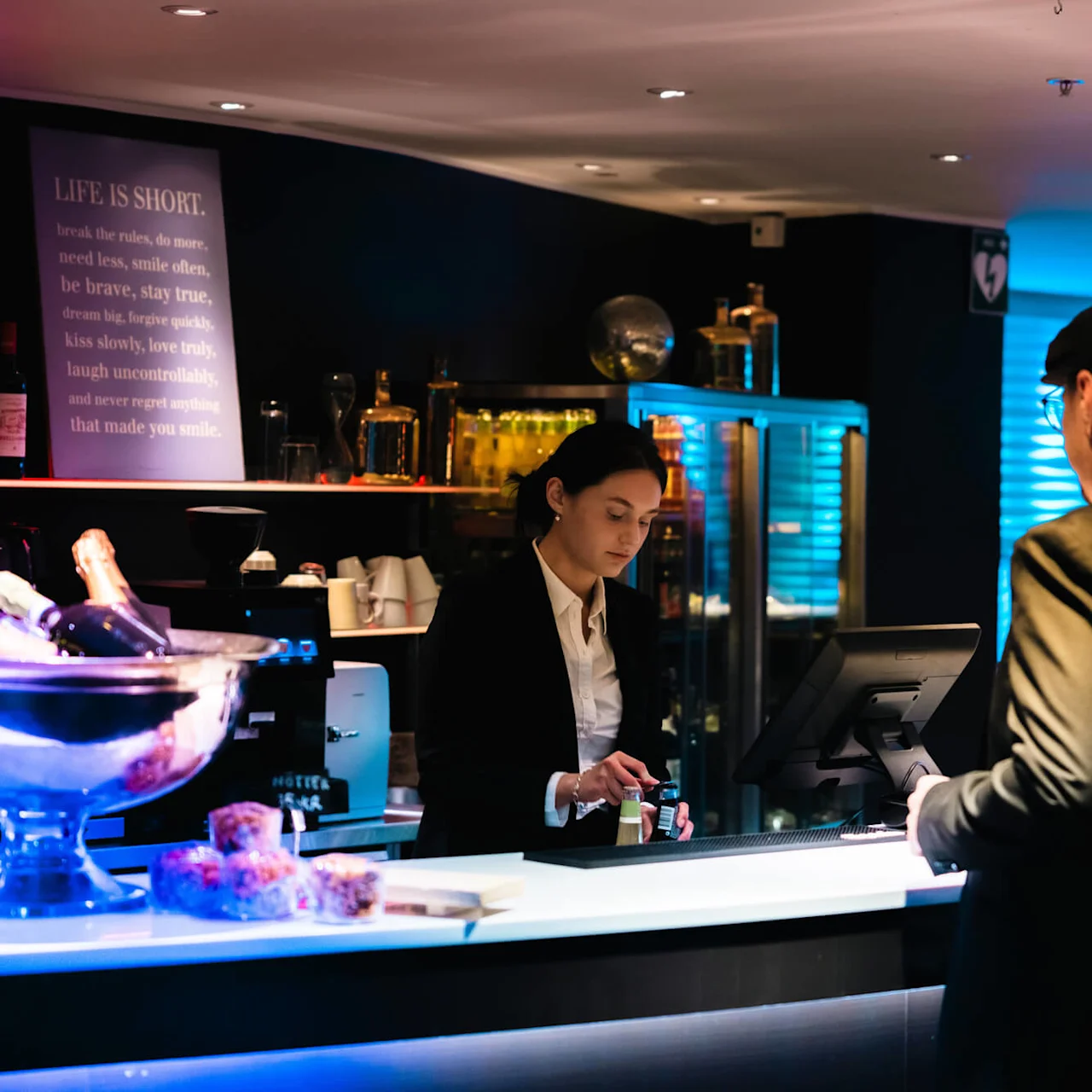 En bartender hanterar betalningar vid en bardisk med drycker på is och snacks förpackade i närheten. Svag belysning framhäver en motivationsskylt med texten: "Life is short..." bakom baren.