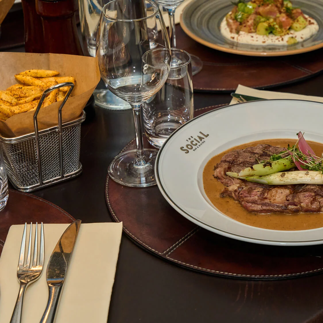 Steak topped with vegetables and greens, served on a sauce-filled white plate labeled "The SOCIAL Marbella," sits on a set dining table with cutlery, crisps, and glassware.