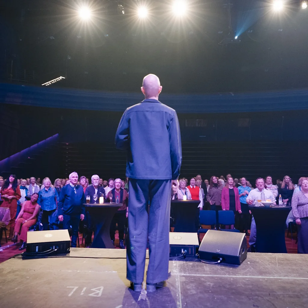 En person på scenen foran et sittende publikum under sterke lys.