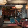 Lyxiga matstolar omger mörka träbord i en elegant upplyst restaurang med omgivande hängande lampor. Glasdörrar med dekorativa mönster skiljer matplatsen från ett mysigt träinrett utrymme.