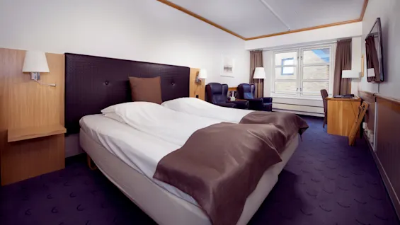En pænt redt seng i et veloplyst hotelværelse med træmøbler, to lænestole og et væghængt TV.