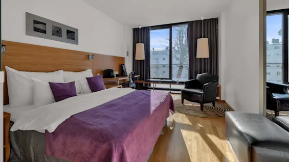 Ett modernt hotellrum med en prydlig säng, stol och stort fönster som släpper in naturligt ljus.