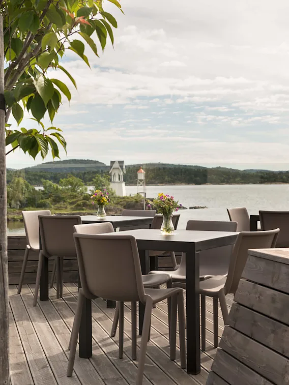 Bord arrangert på en treterrasse har vaser med fargerike blomster; grønne trær og en rolig innsjø med fjerne åser danner den rolige bakgrunnen.