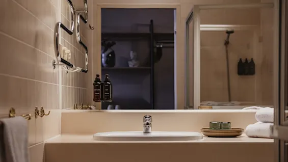 Sink with silver faucet surrounded by beige countertop, soap and lotion bottles, and folded white towels, set against a tiled bathroom wall with a visible shower area.