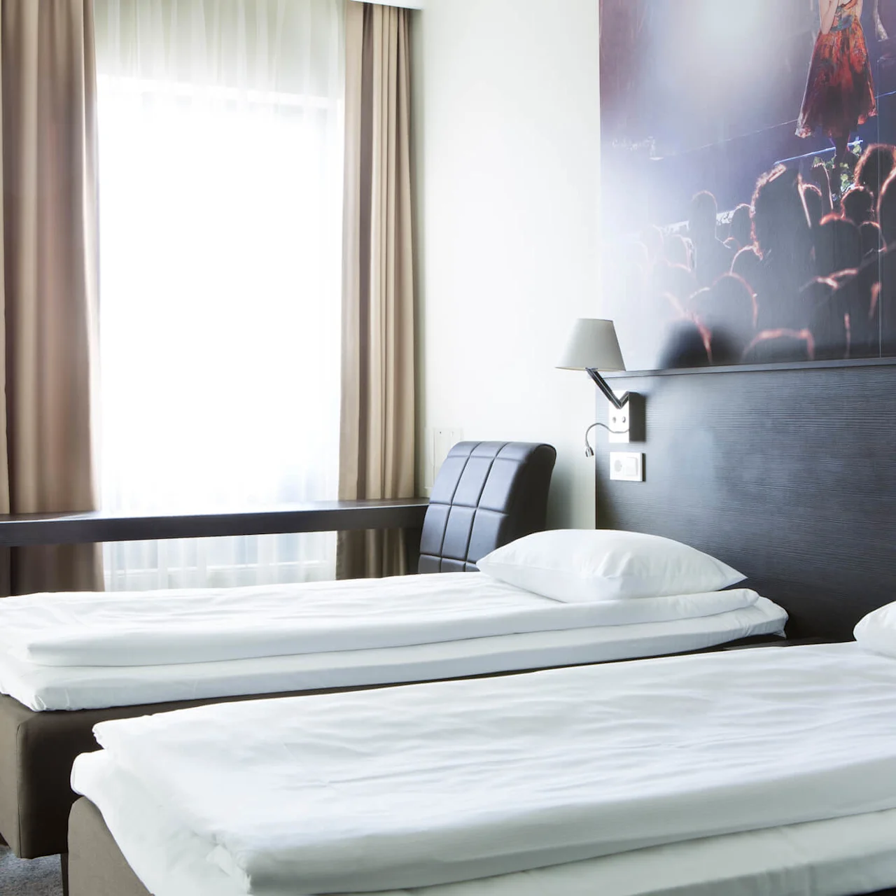 Två prydligt bäddade enkelsängar med vita sängkläder står i ett modernt hotellrum. Ett skrivbord med lampa och stol står bredvid ett fönster med beige gardiner.