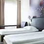 Two neatly made single beds with white linens stand in a modern hotel room. A desk with a lamp and chair sits beside a window with beige curtains.