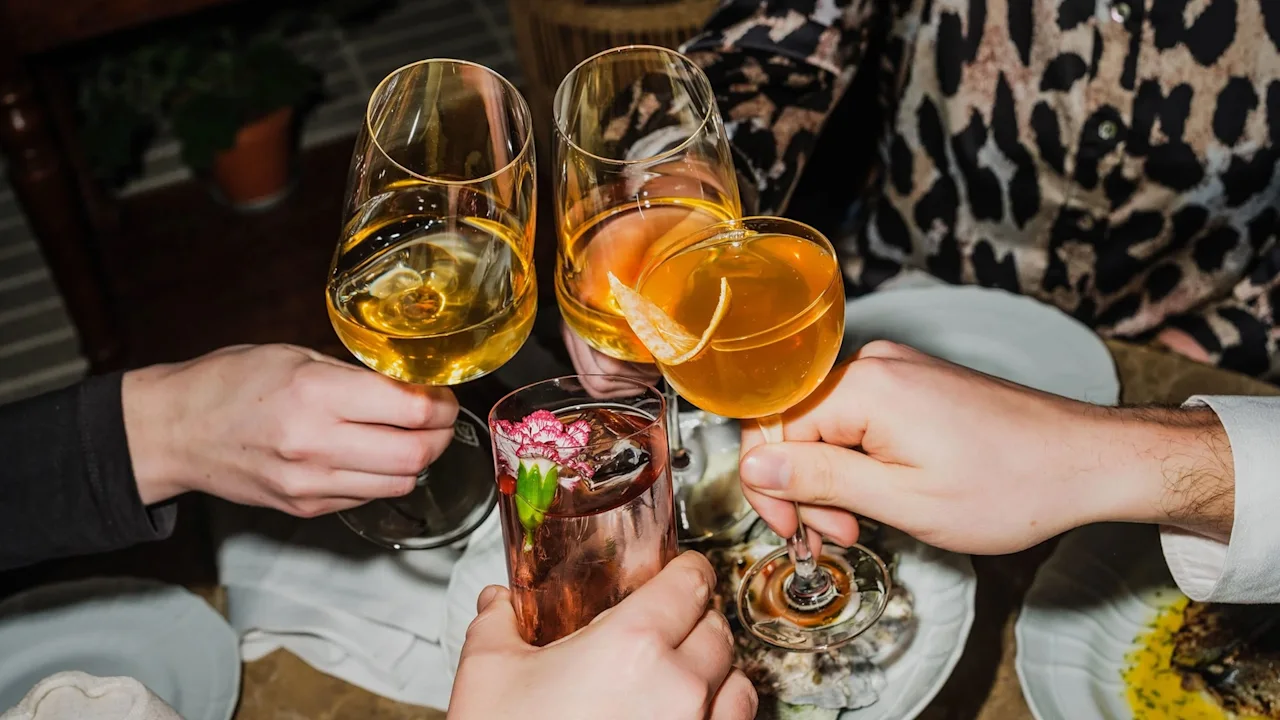 Fire glas klinker sammen i en skål; to med orange cocktails, et med hvidvin, og et med blomsterpynt, i en middagskontekst.