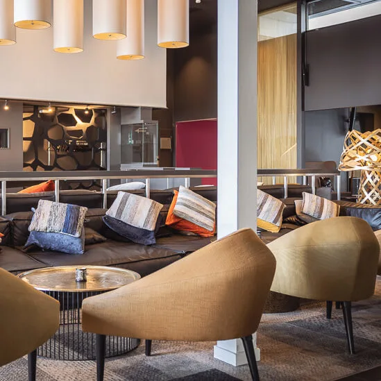 Comfortable armchairs surround a wooden coffee table; behind, a sleek sofa with colorful cushions sits in a modern, warmly lit lounge, featuring geometric wall patterns and pendant lights.