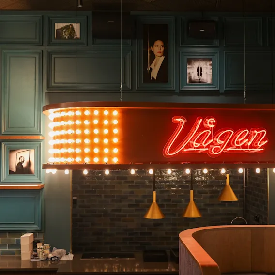 En livlig barinteriör med en neonskylt som lyder Vagen, omgiven av inramade bilder på en turkos vägg.