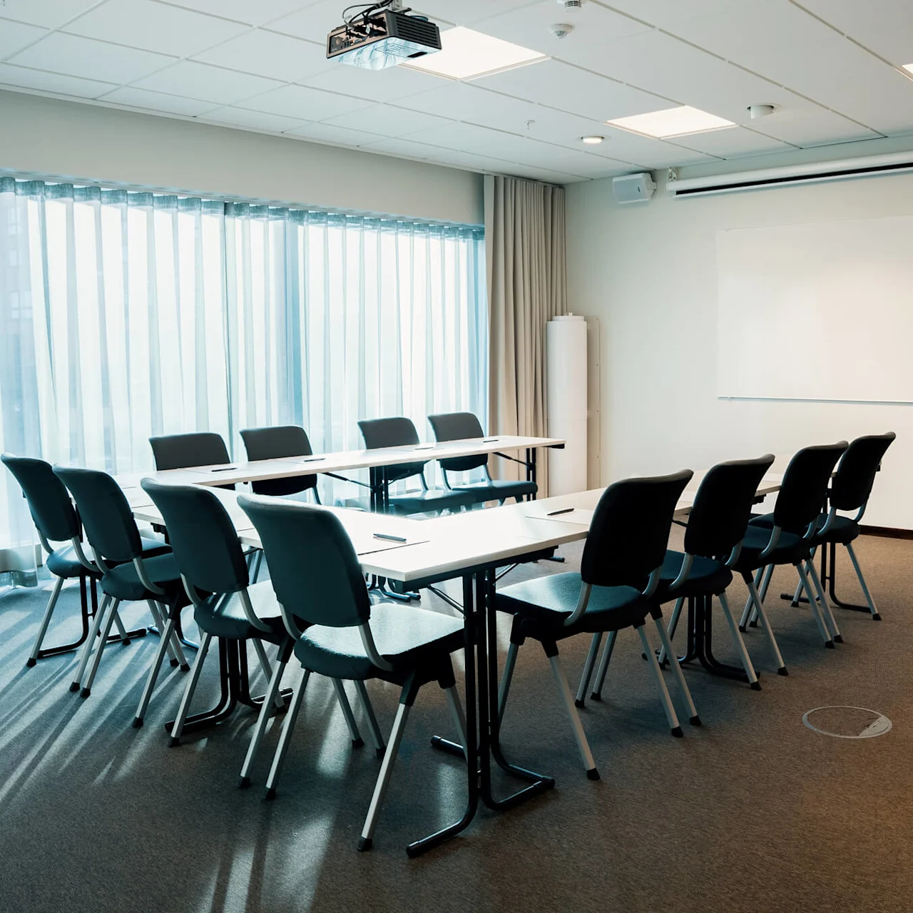 Ett konferensrum har stolar och bord arrangerade i en U-form. En whiteboard och blädderblock står vid väggen. Mjukt naturligt ljus kommer in genom stora fönster med gardiner.