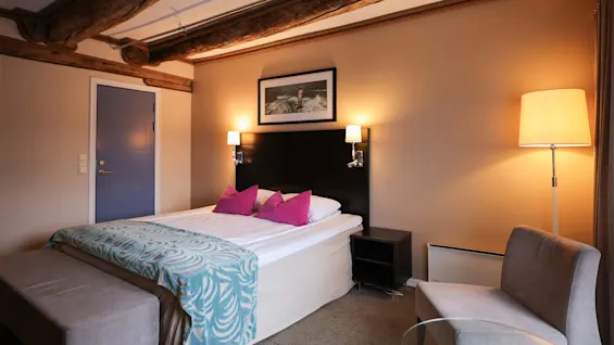 A bed with pink pillows and a teal patterned throw sits in a cozy room featuring exposed wooden beams, a blue door, a lamp, and a framed photo of a lighthouse.