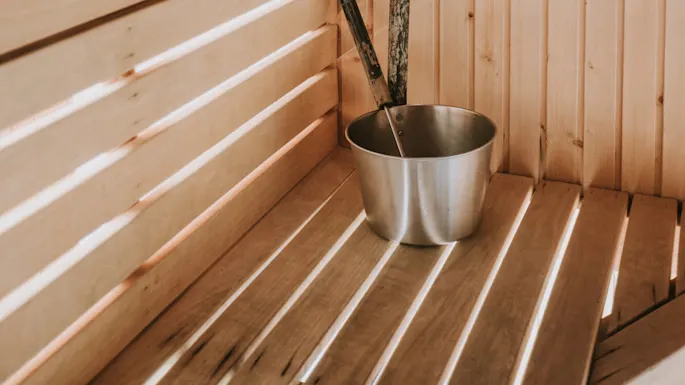 En trebadstu med en metallbøtte og øse på benkene, opplyst av skrått lys som skaper en varm atmosfære.