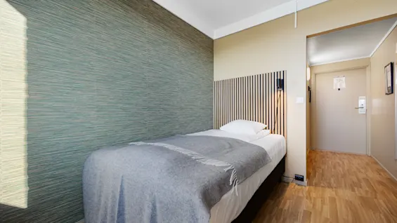 Et koselig hotellrom med en enkeltseng, grønn tapetvegg og lyst tregulv.