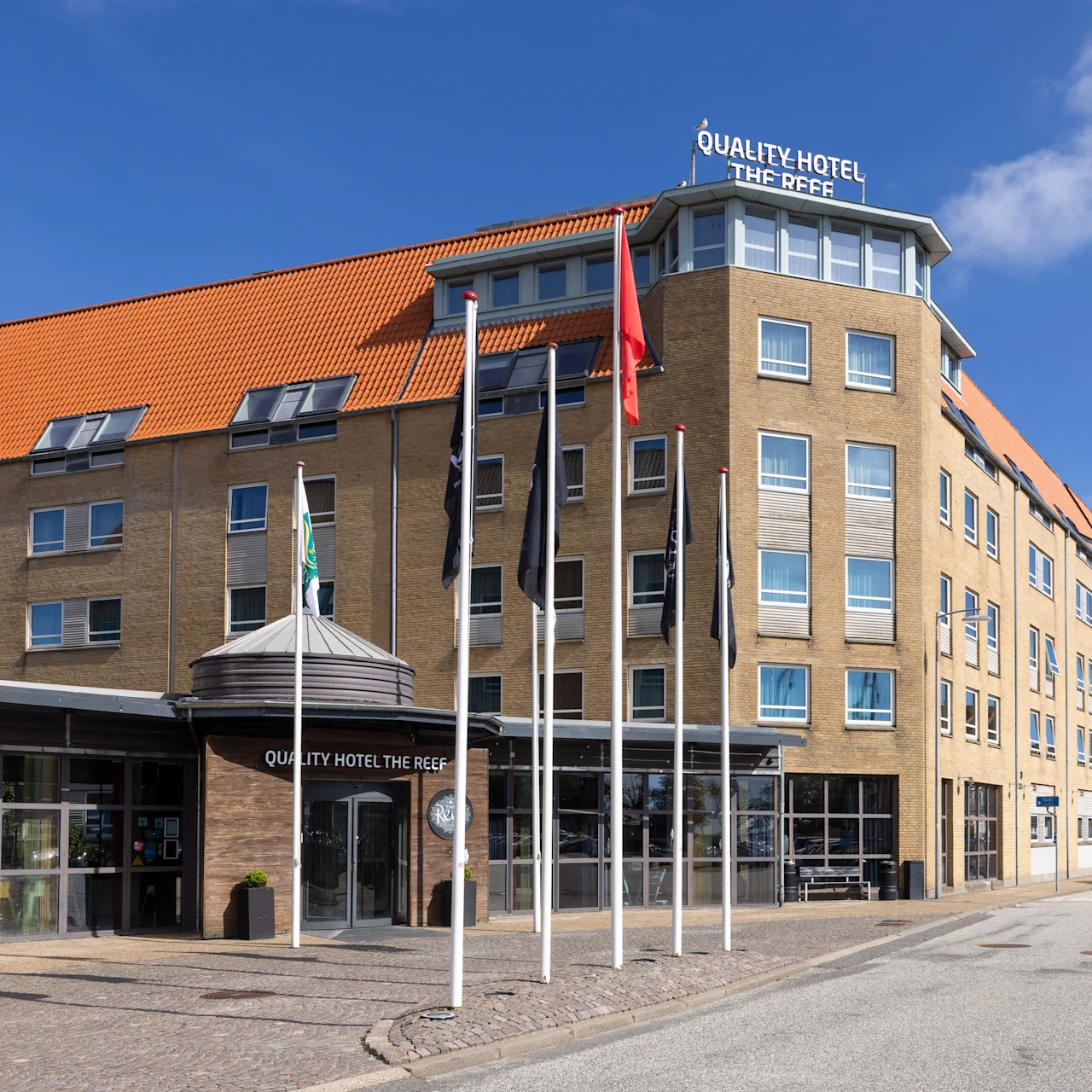 A multi-story hotel with red roof tiles stands on a sunny street, displaying signs reading "QUALITY HOTEL THE REEF" on its facade. Several flags are positioned in front.