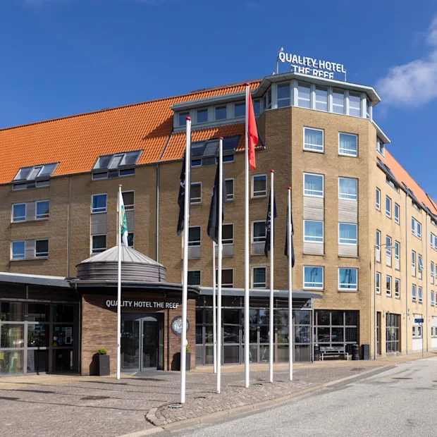 Et fleretagers hotel med rødt tegltag står på en solrig gade, med skilte der viser "QUALITY HOTEL THE REEF" på facaden. Flere flag er placeret foran.