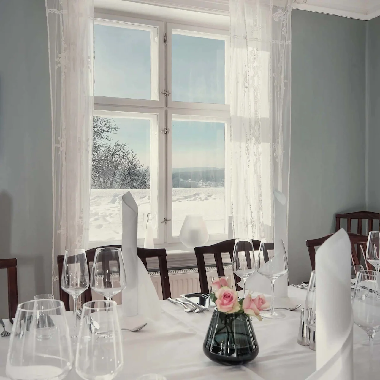 Et spisebord er elegant dækket med glas og en lille vase med lyserøde roser. Sollys strømmer gennem et stort vindue og afslører et sneklædt landskab udenfor.