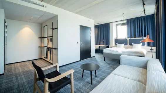 Hotellihuoneessa on parivuode ja oleskelualue, jossa on sohva ja tuoli, aseteltuna moderniin minimalistiseen tyyliin. Huoneessa on siniset verhot ja ikkuna vieressä työpöytä.
