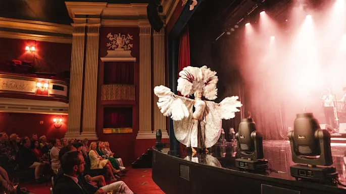 A performer wearing an elaborate feather costume dances on a stage, surrounded by elegant theater interiors, as an attentive audience watches from red velvet seats.