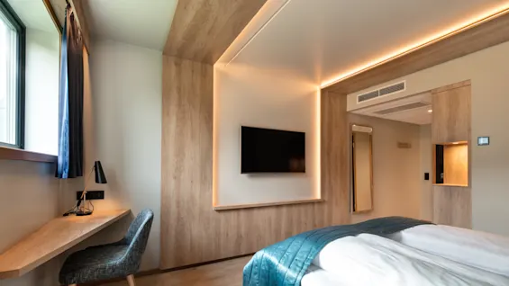 Ett prydligt hotellrum med en säng och ett blått överkast, en väggmonterad TV och ett skrivbord i trä nära ett fönster. Varm belysning förstärker den mysiga atmosfären.