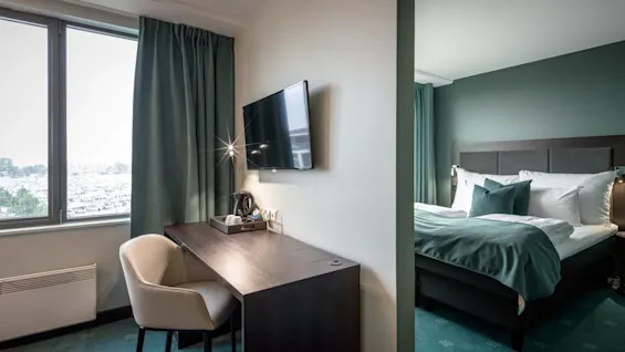 Skrivebord med stol og TV, viser halvdelen af et hotelværelse med seng og grøn indretning. Et vindue med gardiner på venstre side lukker udendørs lys ind.