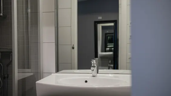 A white sink with a silver faucet sits in a tiled bathroom. A mirror above reflects another room, showing multiple mirror reflections, creating a tunnel effect.