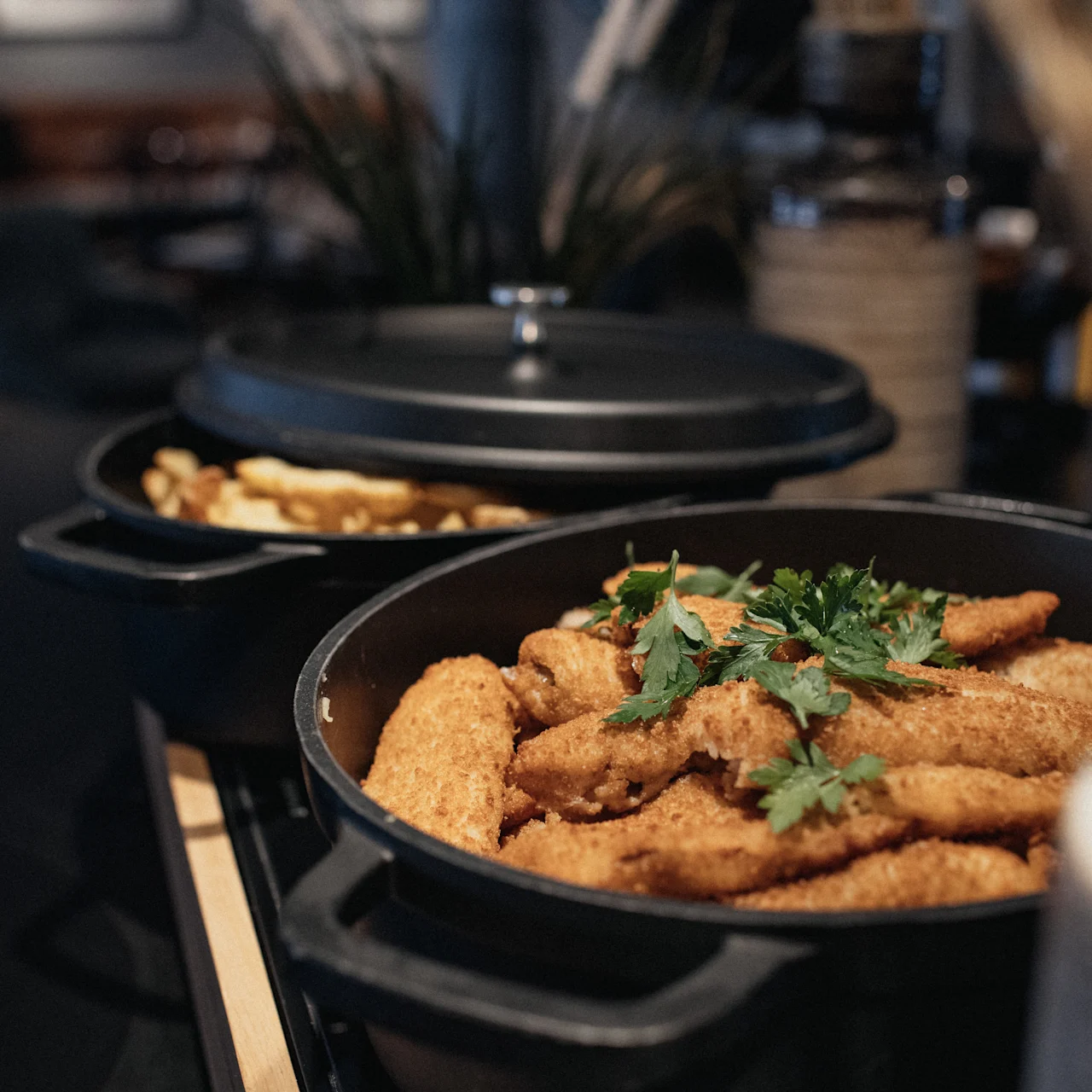 Crispy breaded chicken pieces are garnished with parsley and served in a dark pot on a table. Another pot with fries is nearby, in a warmly lit dining setting.