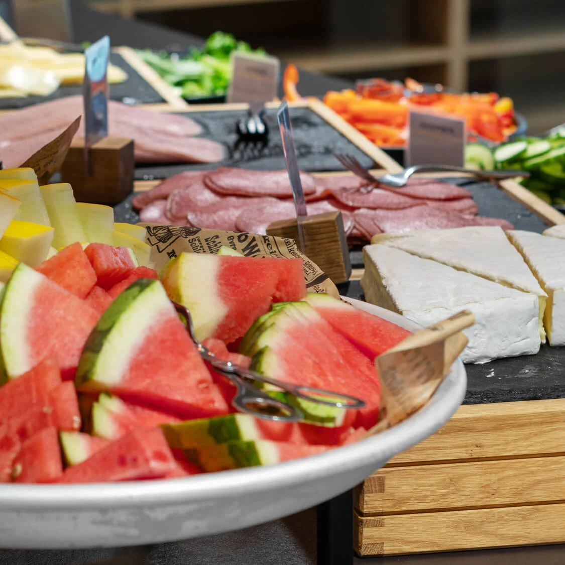 Skivor av olika frukter som vattenmelon, melon och ananas är arrangerade på ett serveringsfat tillsammans med charkuterier och ost på ett buffébord.