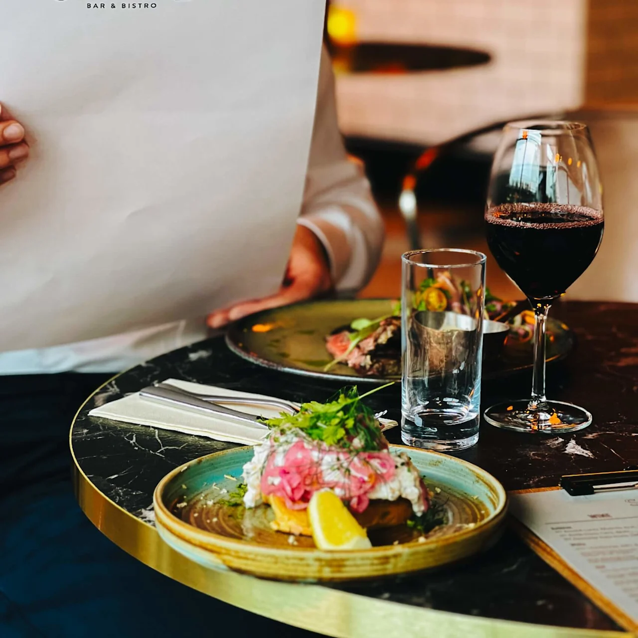 A person holds a menu from "The Social Bar & Bistro” while seated at a table with a plate of food, a glass of wine, and a dimly lit restaurant backdrop.