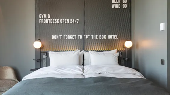 A neatly made double bed with white pillows and a dark blanket is flanked by wall lamps. Text: "GYM & FRONTDESK OPEN 24/7," "DON'T FORGET TO '#' THE BOX HOTEL," "BEER 69," "WINE 99."