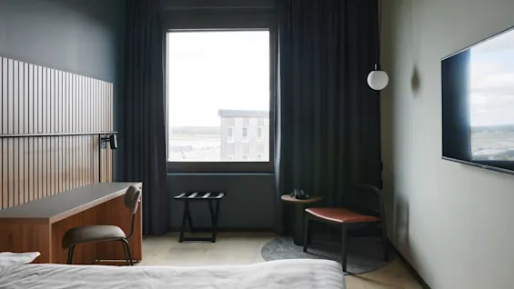 A neatly made bed faces a wooden desk and upholstered chair by a large window with closed curtains, beside a TV and a small seating area with a round table.