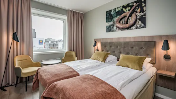 Et hotellrom med to senger, to gule stoler og et vindu med utsikt over en by. Et stort abstrakt kunstverk henger over sengene.