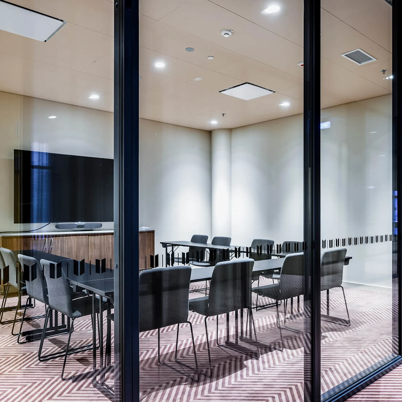 A modern conference room with a large TV on the wall, several chairs around tables, enclosed by glass walls, and featuring a patterned carpet.