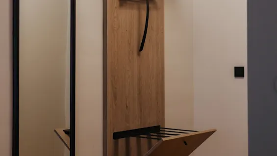 Wall-mounted wooden rack holds hangers and a pull-down shelf; adjacent mirror reflects a simple, tidy interior. White walls and wooden flooring provide a neutral backdrop.