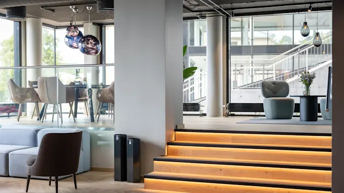 Modern lounge featuring illuminated wooden steps ascending to a seating area with chairs and tables, large windows, and decorative hanging lights, creating a spacious and stylish atmosphere.