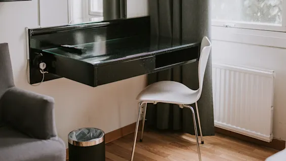 Et sort skrivebord med fjernbetjening i et minimalistisk rum, kombineret med en hvid stol. Omkring er grå gardiner, et vindue, en grå stol og en affaldsspand.