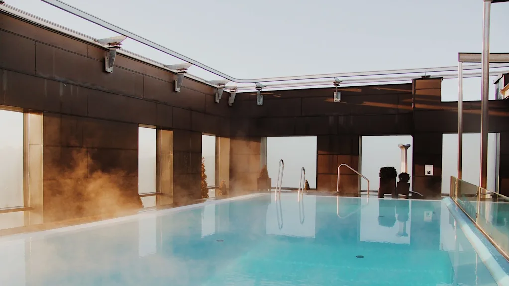 A steaming rooftop pool reflects morning light, surrounded by a modern metal and glass structure, offering a tranquil view of the sky above.