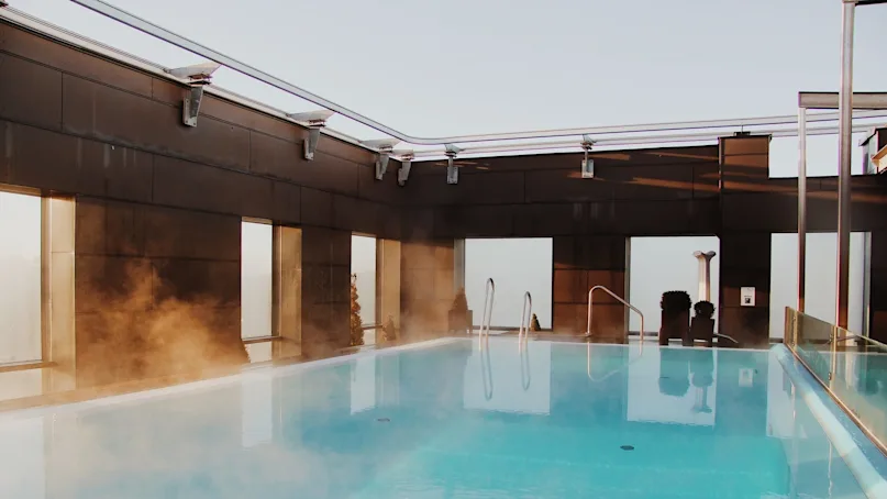A steaming rooftop pool reflects morning light, surrounded by a modern metal and glass structure, offering a tranquil view of the sky above.